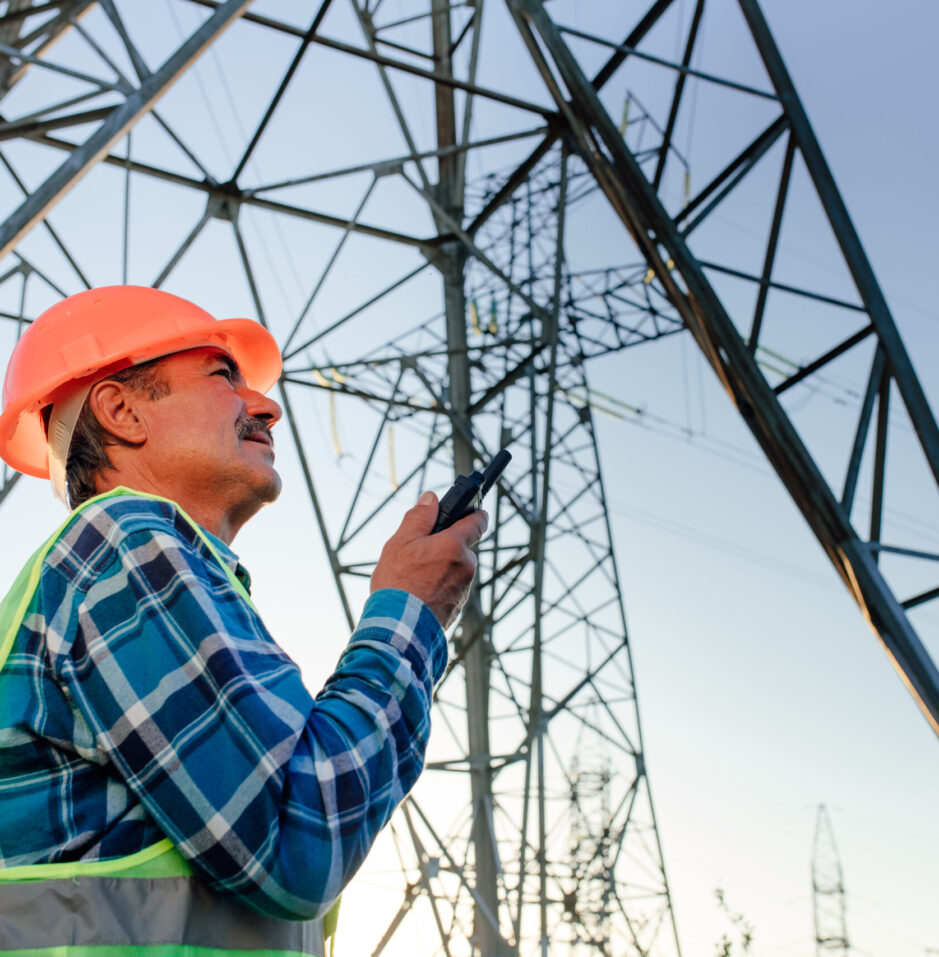 Low angle side view of middle aged male technician in hardhat speaking on walkie talkie while standing in back lit against electric power poles